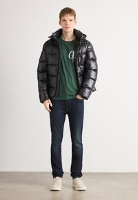 Νεαρός άνδρας όρθιος, φορώντας μαύρη μπουφάν τύπου puffer, πράσινο t-shirt με λογότυπο, σκούρα τζιν και μπεζ αθλητικά παπούτσια, μπροστά σε απλή φόντο.