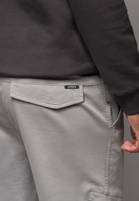 Grijze broek van gladde stof, met een achterzak met klep en een klein zwart label op de tailleband, gecombineerd met een zwart sweatshirt.