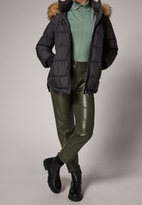 Persona vestida con una chaqueta acolchada negra con capucha de piel, un top de punto verde, pantalones de cuero verde olivo y botas negras de tobillo, de pie con las piernas cruzadas.