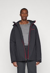 Μαύρο μπουφάν μόνωσης με κόκκινες λεπτομέρειες στη φερμουάρ, ρυθμιζόμενη κουκούλα και πλαϊνές τσέπες. Φοριέται πάνω από ένα γκρι πουκάμισο, με ανθεκτικό εξωτερικό υλικό.