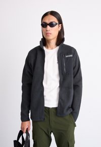 Chaqueta de forro polar gris con bolsillo en el pecho con cremallera, llevada sobre una camisa blanca. La persona sostiene un bolso negro y lleva gafas de sol negras. Pantalones verde oliva.