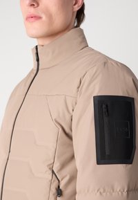 Beige puffjacka med hög krage, svart dragkedja och texturerad axelficka med dragkedjestängning och logodetalj.