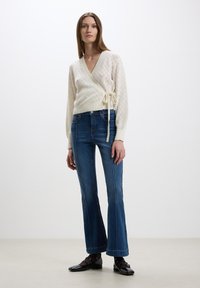 Maglione avvolgente bianco lavorato a maglia con motivo a diamante, cintrato in vita, abbinato a jeans blu a zampa d'elefante e scarpe nere basse.