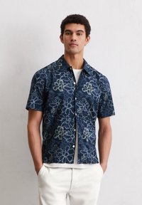 Kortemouw navyblauw shirt met wit bloemenpatroon, knoopsluiting aan de voorkant en een relaxed fit. Draagt over een wit T-shirt.