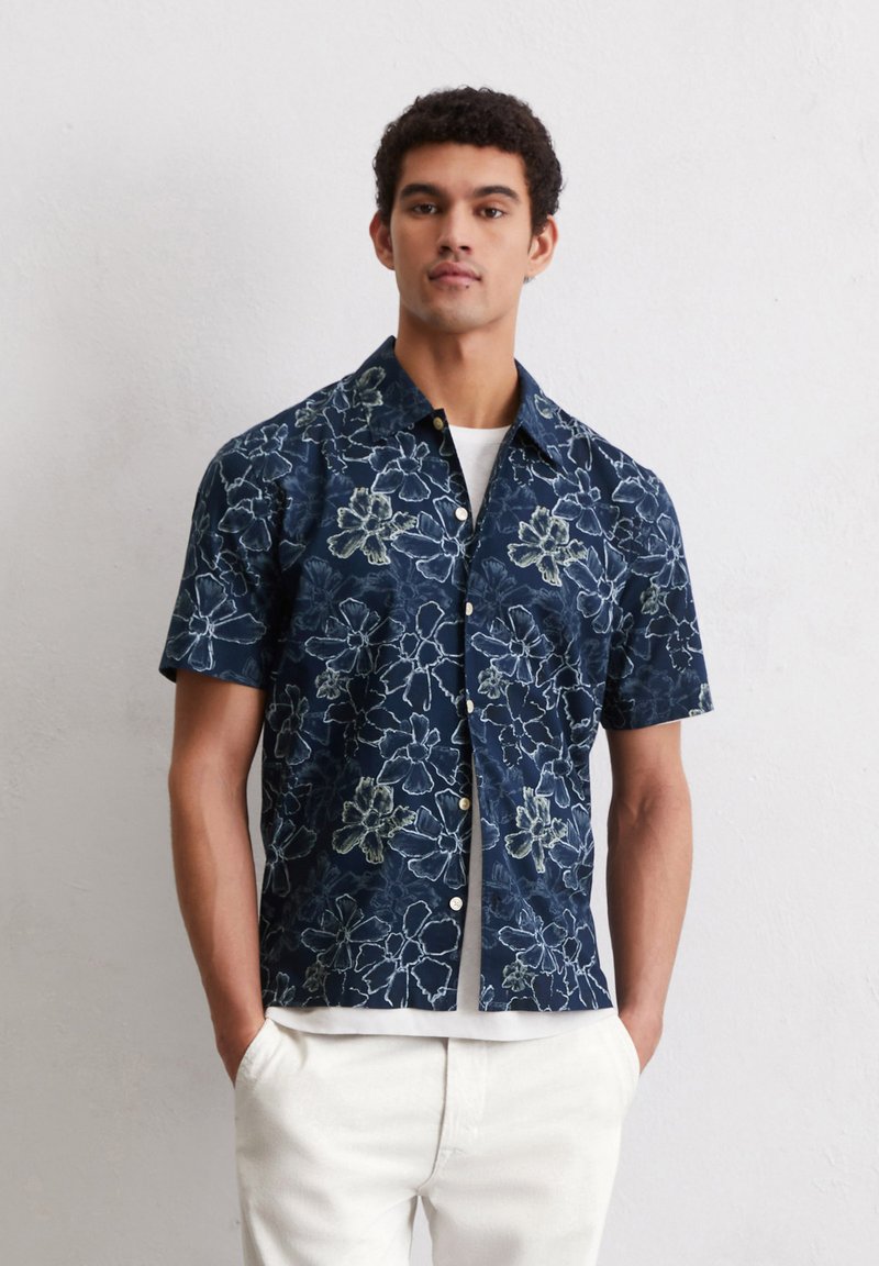 Kortemouw navyblauw shirt met wit bloemenpatroon, knoopsluiting aan de voorkant en een relaxed fit. Draagt over een wit T-shirt.