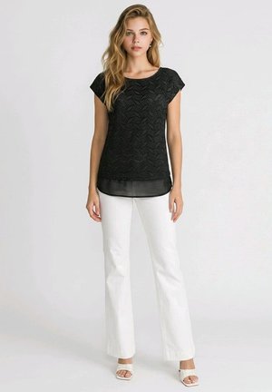Mujer con cabello rubio largo que lleva una blusa negra texturizada de manga corta con dobladillo transparente, pantalones blancos y tacones blancos abiertos en los dedos, de pie frente a un fondo simple.