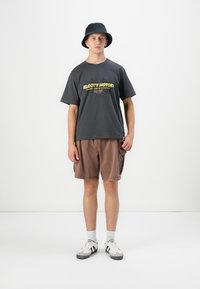 Ung mand iført sort bucket hat, mørk grafisk T-shirt, brune cargo shorts, hvide sokker og hvide sneakers, står mod en hvid baggrund.