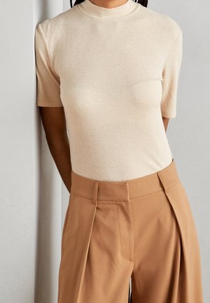 Personne portant un haut beige côtelé à manches courtes, rentré dans un pantalon plissé camel à taille haute, devant un mur clair uni.