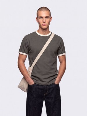 Jeune homme aux cheveux courts portant un t-shirt gris, un jean foncé et un sac bandoulière beige, debout les mains dans les poches.