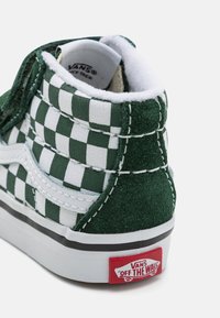Zapatilla de caña alta a cuadros verdes y blancos con textura de ante, que presenta costuras blancas y una etiqueta roja de "Vans Off the Wall" en el talón.
