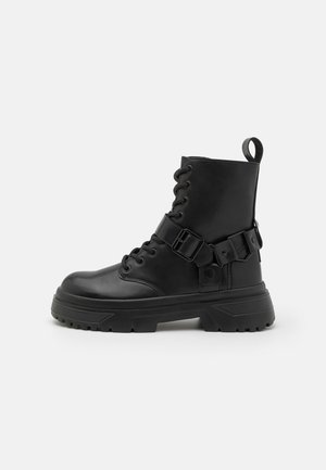 Juodas kovos botas, pagamintas iš lygaus odos, su raišteliais priekyje, iki kulkšnies aukščio, reguliuojamu sagties dirželiu ir storu guminiu padu.