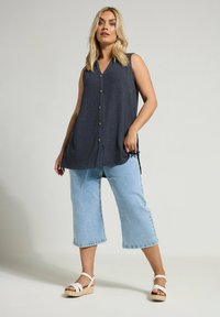 Bezrękawnik w kolorze granatowym z dekoltem w kształcie litery V i guzikami z przodu, zestawiony z jasnoniebieskimi, krótkimi dżinsami oraz białymi sandałami na koturnie.