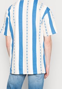 Kortärmad skjorta med blå och beige vertikala ränder, med ett mönster av små pilar. Bomullstyg, avslappnad passform, rundad halsringning.