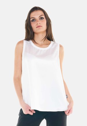 Jeune femme aux longs cheveux bruns portant un haut blanc sans manches et un pantalon noir sur un fond clair uni.