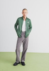 Casaco leve e verde com fecho éclair sobre uma camisa branca, combinado com calças de veludo cotelê cinzas e sapatos pretos. Design simples com acentos mínimos.