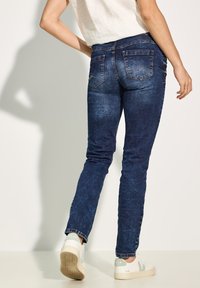 Sleek, ciemnoniebieskie jeansy, wykonane z denimu, z tylnymi kieszeniami ozdobionymi przeszyciami. W połączeniu z białymi trampkami i jasną bluzką.