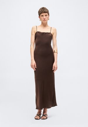 Kvinde med kort hår iført en brun satin maxikjole uden ærmer og sorte sandaler, accessoriseret med gyldne øreringe og en manchetarmbånd.