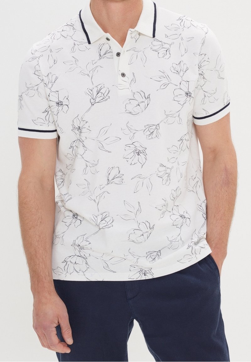 Camisa polo blanca con arte lineal floral negro, abertura de tres botones y ribete en azul marino en el cuello y las mangas. Material de algodón suave con mangas cortas.