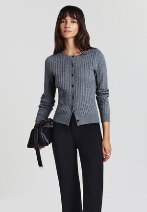 Frau in einem grauen, kabelgestrickten, geknöpften Cardigan, schwarzen Hosen, die eine schwarze Lederhandtasche mit einem langen Tragegurt hält und vor einem schlichten Hintergrund steht.