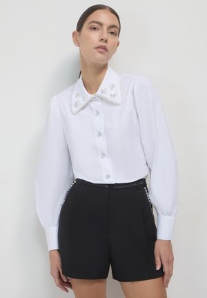 White button-up shirt with a large embellished collar featuring pearls and flowers, paired with black tailored shorts. Smooth fabric, long sleeves.