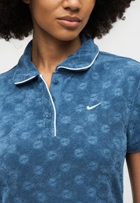 Γυναίκα που φοράει μπλε πόλο πουκάμισο με υφή και λευκή λεπτομέρεια, με μικρό λογότυπο Nike στο αριστερό στήθος, εμφανιζόμενη από τους ώμους μέχρι τα χείλη.