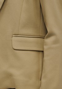 Gros plan sur une manche de blazer beige et une poche à rabat avec des coutures visibles et la texture du tissu.