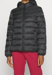 Svart vadderad jacka med huva och framdragkedja, bärs med röda byxor, delvis synlig person som står mot neutral bakgrund.