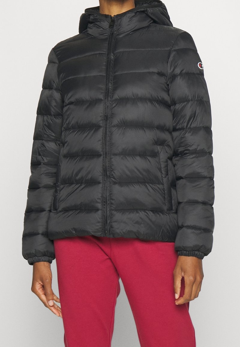 Svart vadderad jacka med huva och framdragkedja, bärs med röda byxor, delvis synlig person som står mot neutral bakgrund.