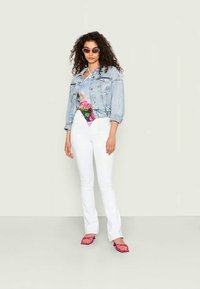 Casaco de ganga, bodysuit floral com detalhes em rosa, jeans brancos à boca de sino e sandálias rosas. Modelo com cabelo encaracolado e óculos de sol redondos.