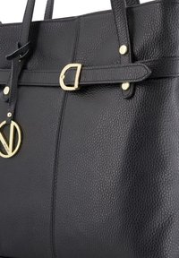 Sac fourre-tout en cuir noir avec une texture granuleuse, des accents matériels dorés, un pendentif logo distinctif et des détails ceinturés à l'avant.