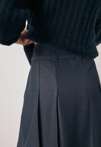 Tume sinine volditud seelik, millel on istuv vöökoht vööaasadega ja peened õmblused. Valmistatud siledast, tekstureeritud kangast.