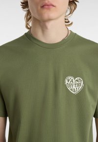 Camiseta de manga corta color verde oliva hecha de algodón, con un pequeño logo de un corazón blanco que presenta el texto "VANS OFF THE WALL" en el área del pecho izquierdo.