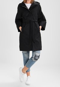 Černý trench coat se zapínáním na zip a kapucí, s širokými rukávy. Vybavený lehkými modrými džíny a bílými teniskami.