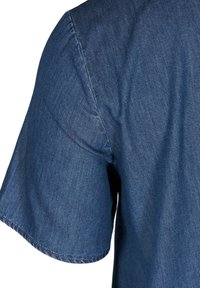 Chemise à manches courtes en tissu denim bleu, montrant la partie supérieure du bras et la zone de l'épaule avec des coutures visibles et une texture.