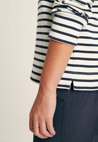 Langärmliges gestreiftes Oberteil in Weiß und Marineblau, mit Rundhalsausschnitt, gerippten Bündchen und einem seitlichen Schlitz am Saum. Hände mit Ringen sichtbar.