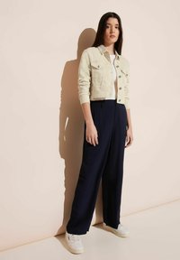 Ljusbeige kort denimjacka, vit skjorta, marinblå vida byxor och vita sneakers, med knappar och fickor synliga. Släta texturer.