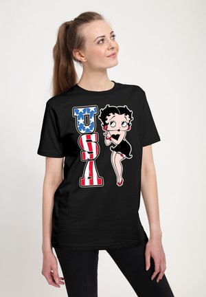 T-shirt noir présentant un personnage de dessin animé, "Betty Boop," avec des lettres "USA" rouges, blanches et bleues, des motifs d'étoiles et des escarpins rouges.