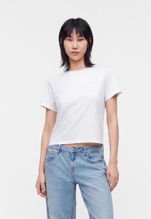 Person står mod en ensfarvet baggrund iført en hvid kortærmet t-shirt og lyseblå jeans, med et neutralt ansigtsudtryk.