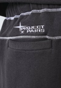 Primer plano de un bolsillo de pantalones de tela oscura con costuras en contraste blancas y el logotipo bordado de "Project X Paris" encima del bolsillo.