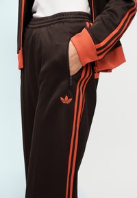 Personne portant un pantalon de survêtement noir et une veste avec des bandes orange, main dans la poche zippée, chemise blanche visible en dessous.
