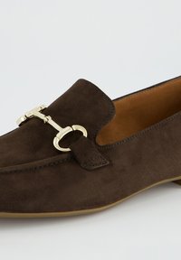 Dunkelbrauner Veloursleder-Slipper mit goldfarbenem Metall-Horsebit-Detail und hellbraunem Innenfutter vor hellem Hintergrund.