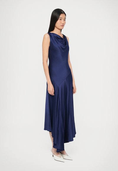 Szatén tengerészkék ruha lezser húzott nyakkivágással, testhezálló felső résszel és bővülő, aszimmetrikus szoknyával, sima textúrával és kecses eséssel. Fehér cipő.