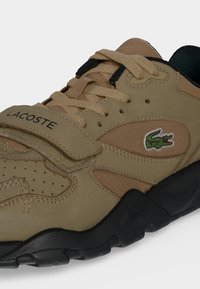 Μπεζ αθλητικό Lacoste με κορδόνια, λουράκι Velcro, πράσινο λογότυπο κροκόδειλου και μαύρη υφή σόλας.