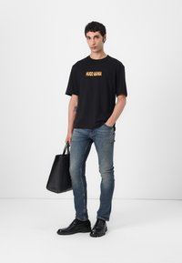 Svart T-skjorte med "HUGO MANIA" i oransje tekst, kombinert med blå jeans, svarte sko og en svart tote bag. Enkelt design med korte ermer.