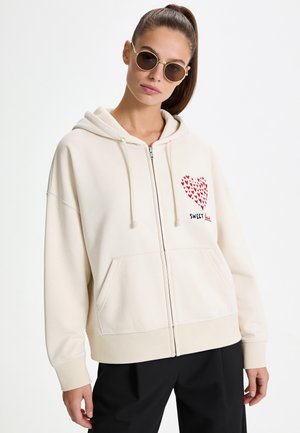 Femme portant des lunettes de soleil rondes et un sweat à capuche crème zippé avec un cœur rouge et le texte "Sweet Heart" sur la poitrine, associé à un pantalon noir.