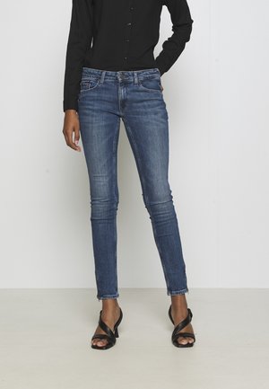 Woman wearing dark blue skinny jeans, black button-up shirt, and black heeled sandals standing against plain light background.