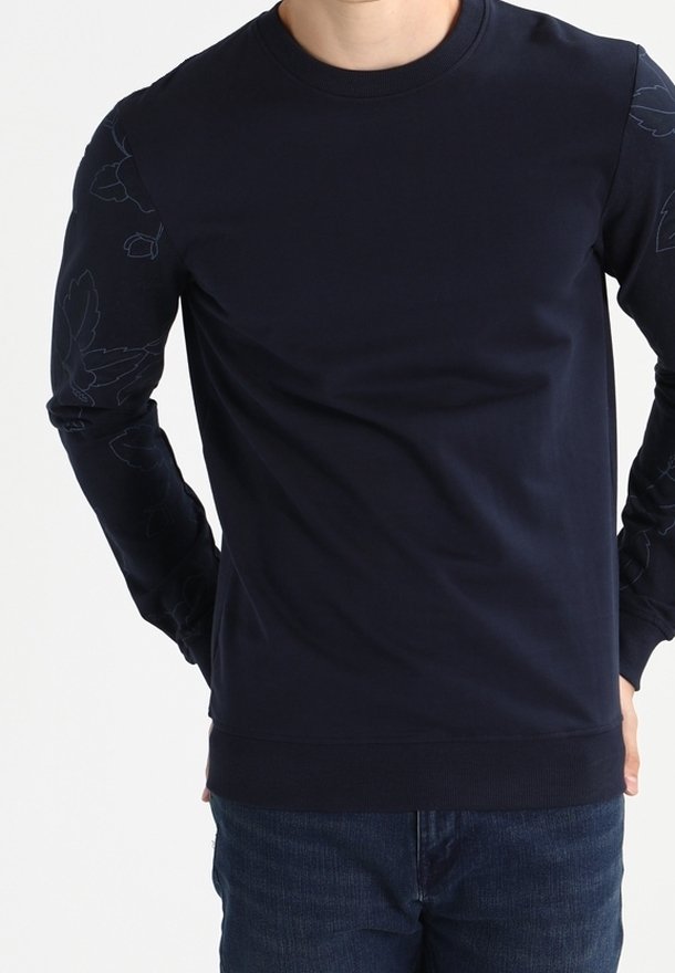 Homme portant un sweat-shirt bleu marine foncé avec une broderie florale subtile sur les manches et un jean bleu, debout les mains derrière le dos.
