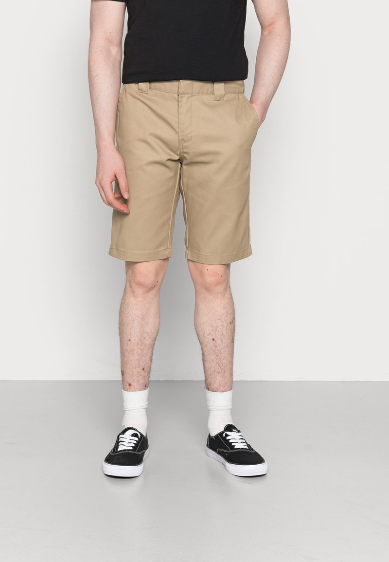 Homme portant un short beige jusqu'aux genoux, un t-shirt noir, des chaussettes blanches et des baskets noires, debout avec une main dans la poche.