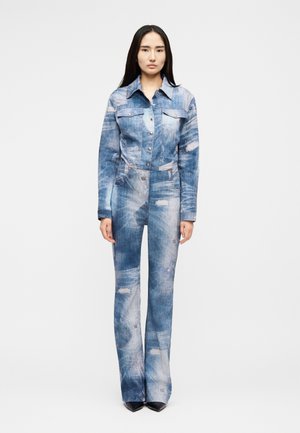 Egy nő áll, visel egy összeillő, kifakult kék farmer dzsekit és farmernadrágot kopott részletekkel, szemben áll egy egyszínű fehér háttér előtt.