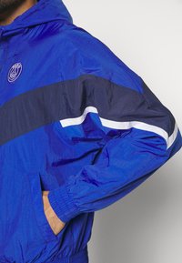 Man som bär en blå Paris Saint-Germain vindjacka med handen i fickan, med marinblå och vita ränder på ärmen.